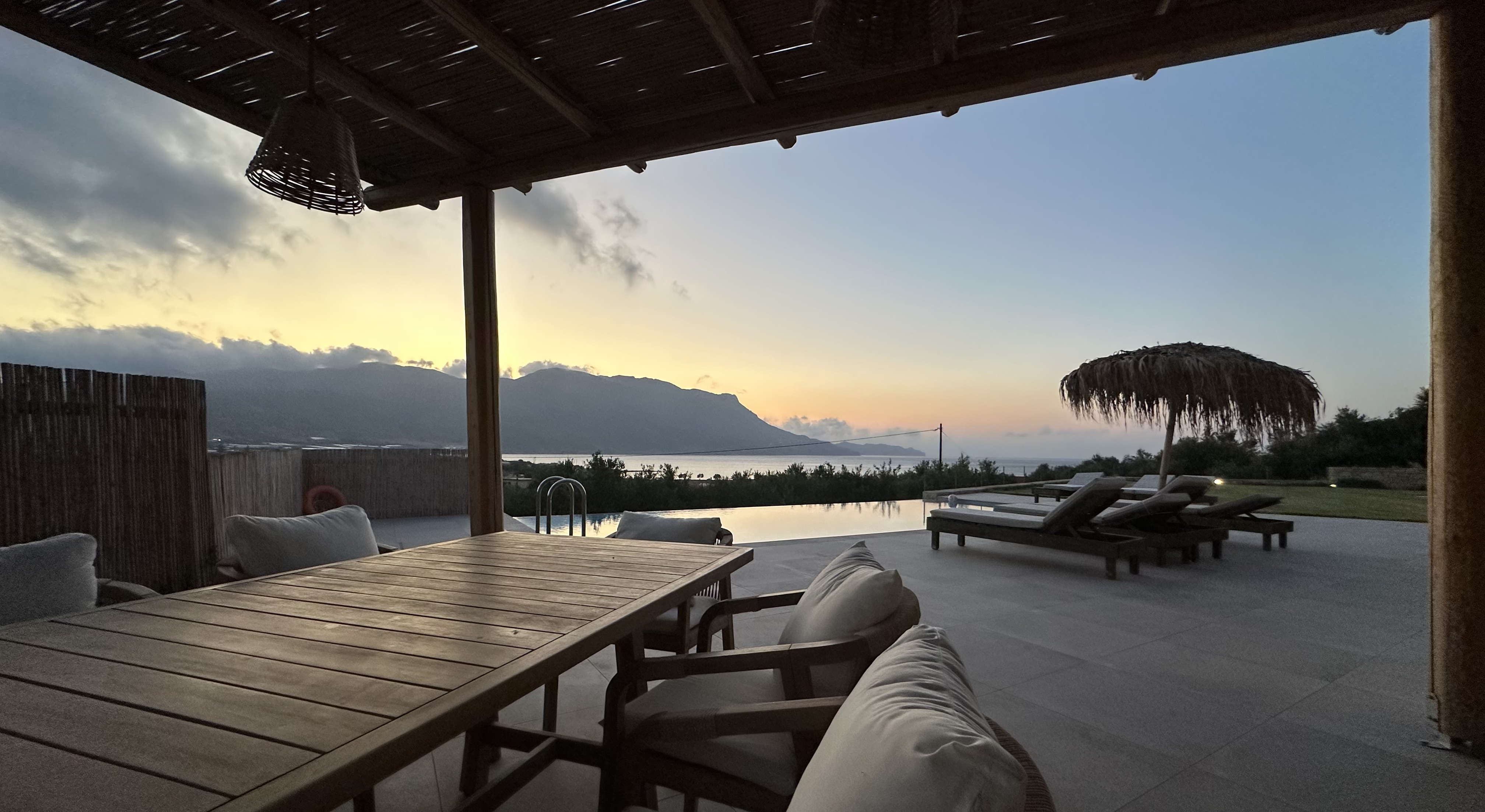 Pergola terrace with dining table looking out over infinity pool and bay at sunset, NUSA Falasarna villa, Crete