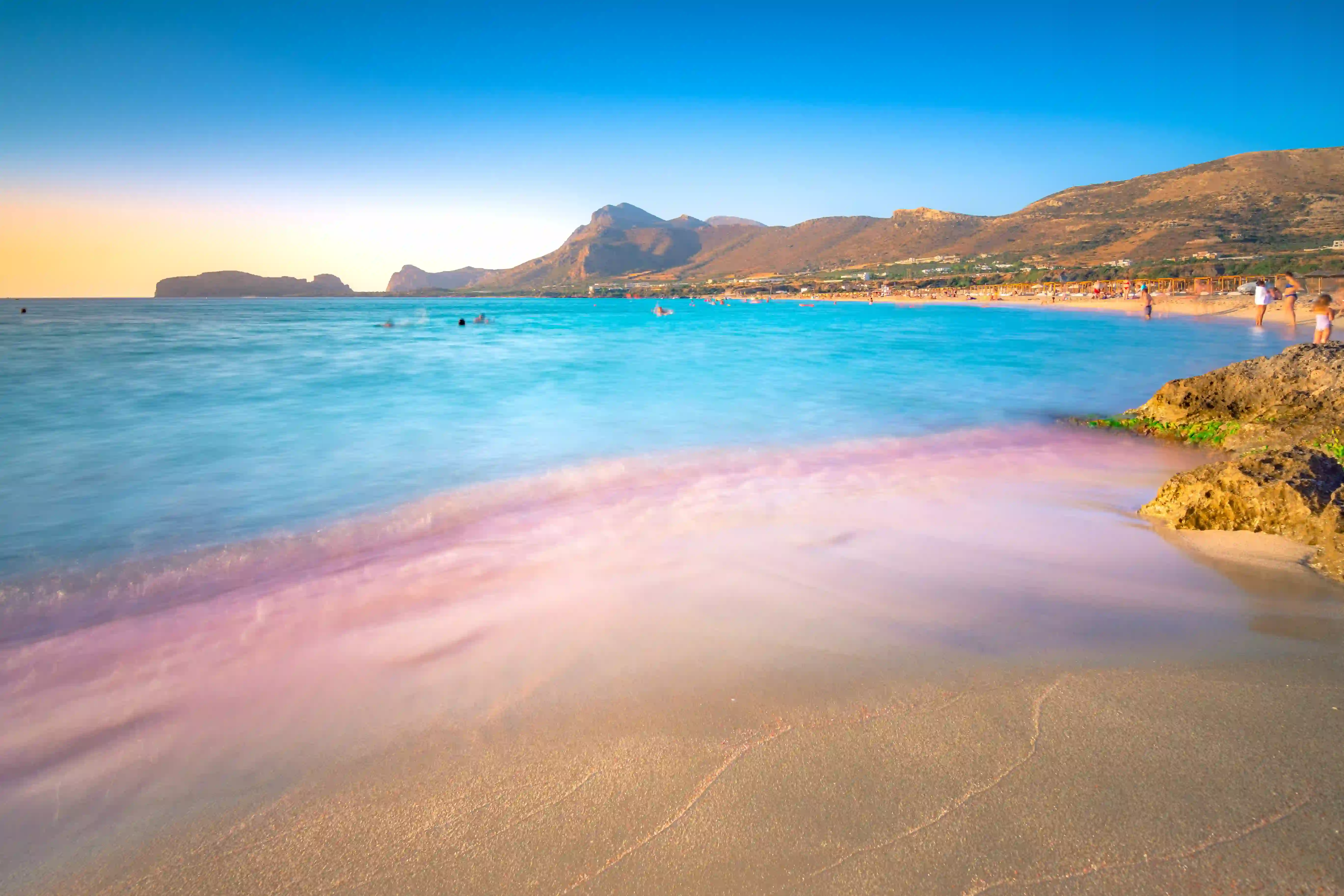 Falassarna Beach at sunset with pink-gold light reflecting on shallow waters and mountains in background, Crete